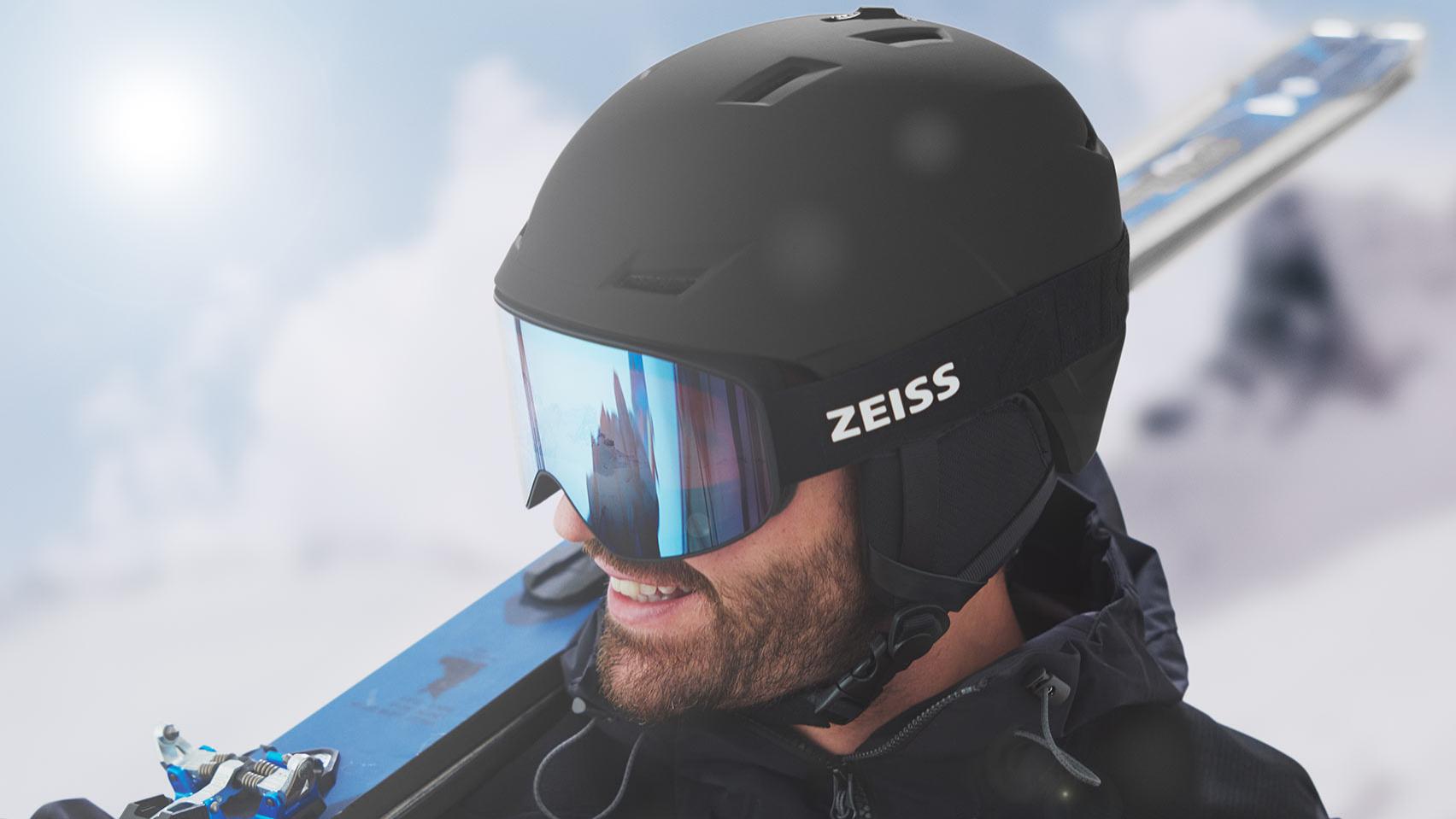 Мъж носи черна каска със сини огледални скиорски очила ZEISS, а през дясното рамо има ски.